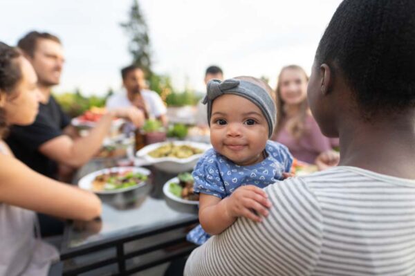 A multiethnic group of young friends enjoy good food and conversation together on a terrace outside on a summer evening. The focus is on an adorable young girl who's smiling at the camera while mom holds her.