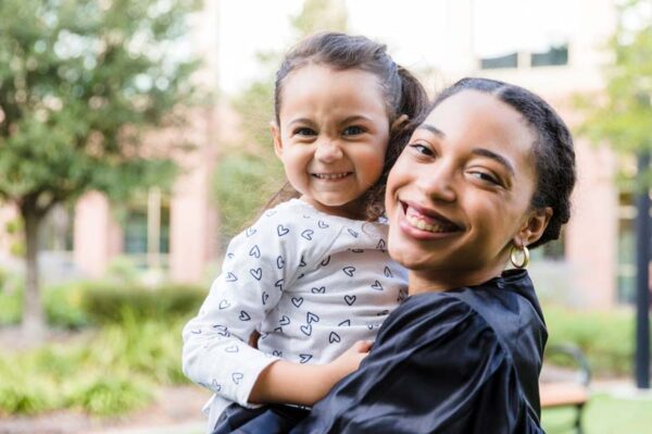Young female college graduate holds her daughter after graduating. The woman and her daughter are smiling at the camera.